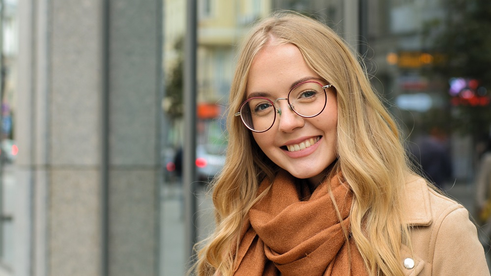 Blonde Woman in Glasses and Scarf Smiling Outdoors - Mount Carmel Eye Doctor Young blonde woman wearing round glasses and a brown scarf smiles warmly while standing outside in an urban setting - Mount Carmel Eye Doctor.