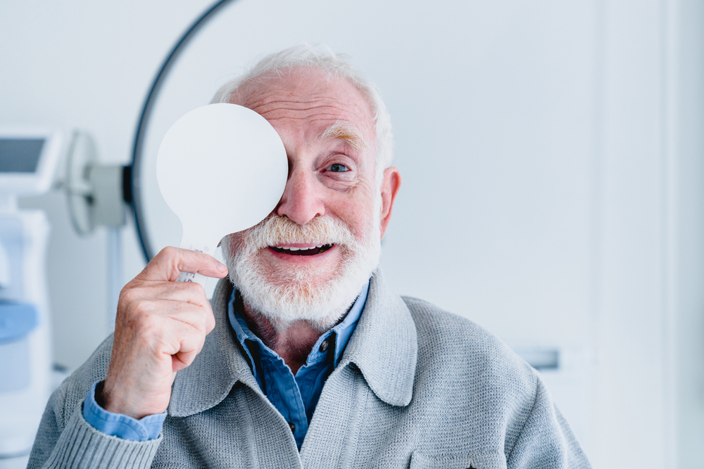 Elderly Man Smiling During Vision Test A cheerful senior man with white hair and beard covers one eye with a testing paddle during an eye exam in a bright clinic.