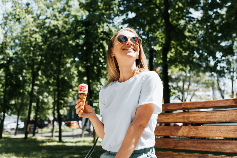 Woman Enjoying Ice Cream Outdoors - Eye Doctor in Kingsport, TN Smiling woman in sunglasses and a white t-shirt enjoys an ice cream cone while sitting on a park bench in the sunshine - Eye Doctor in Kingsport, TN.