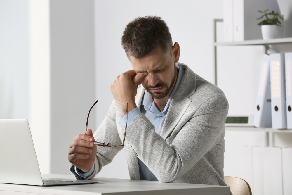 Man Experiencing Eye Strain at Work A man in a light gray blazer sits at a desk with a laptop, rubbing his eyes while holding eyeglasses, showing signs of digital eye strain.