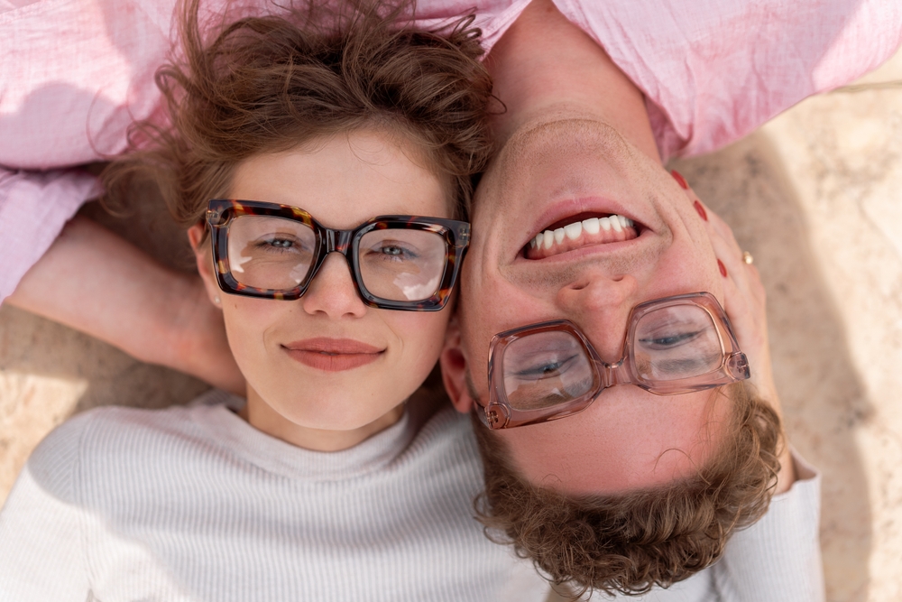 Stylish Vision for Two - McMillin Eyecare A smiling couple wearing fashionable glasses lies on the ground, looking up. - McMillin Eyecare