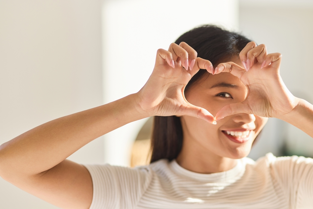 Smiling Woman Making Heart Shape with Hands Around Eye A cheerful woman forms a heart shape with her hands around one eye, symbolizing love and care for healthy vision.