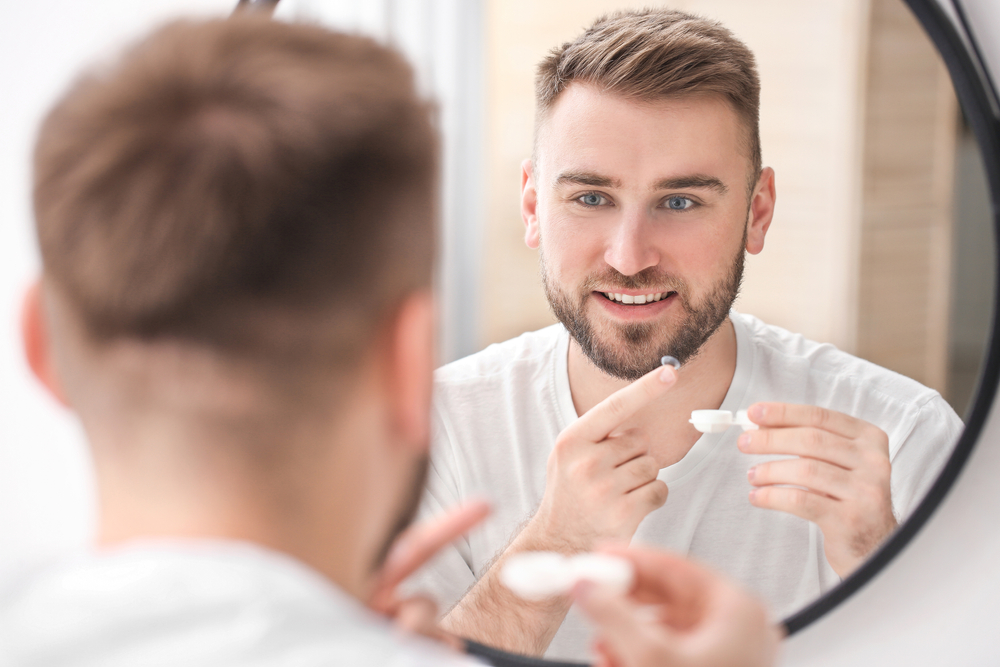 Effortless Vision with Contacts - McMillin Eyecare A smiling man looks in the mirror while preparing to put in a contact lens. - McMillin Eyecare