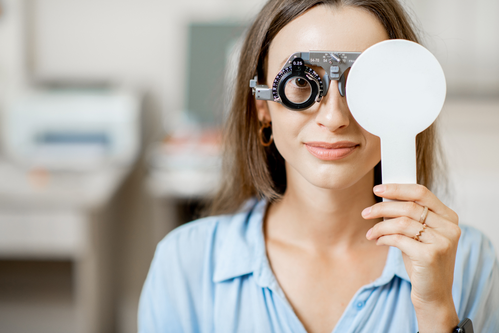 Woman Holding Occluder During Eye Test - Eye Exam Kingsport TN Young woman in a blue shirt holds an occluder over one eye while wearing a trial frame for an eye test - Eye Exam Kingsport TN.