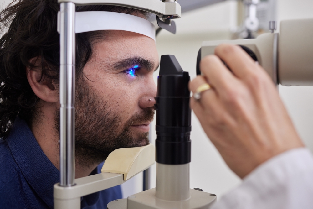 Man Undergoing Slit Lamp Eye Examination Close-up of a man with dark hair and beard receiving a slit lamp eye exam, with blue diagnostic light reflecting in his eye.