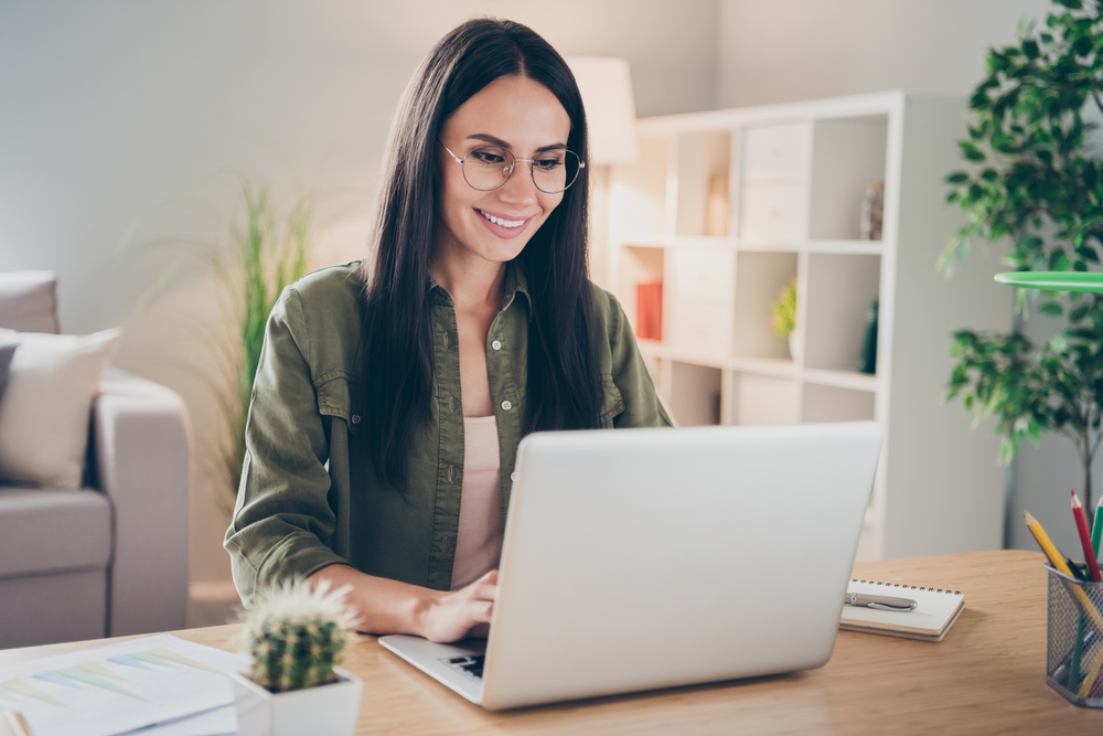 Woman Working on Laptop with Glasses A woman wearing glasses smiles while working on a laptop at home, surrounded by bookshelves, plants, and office supplies.