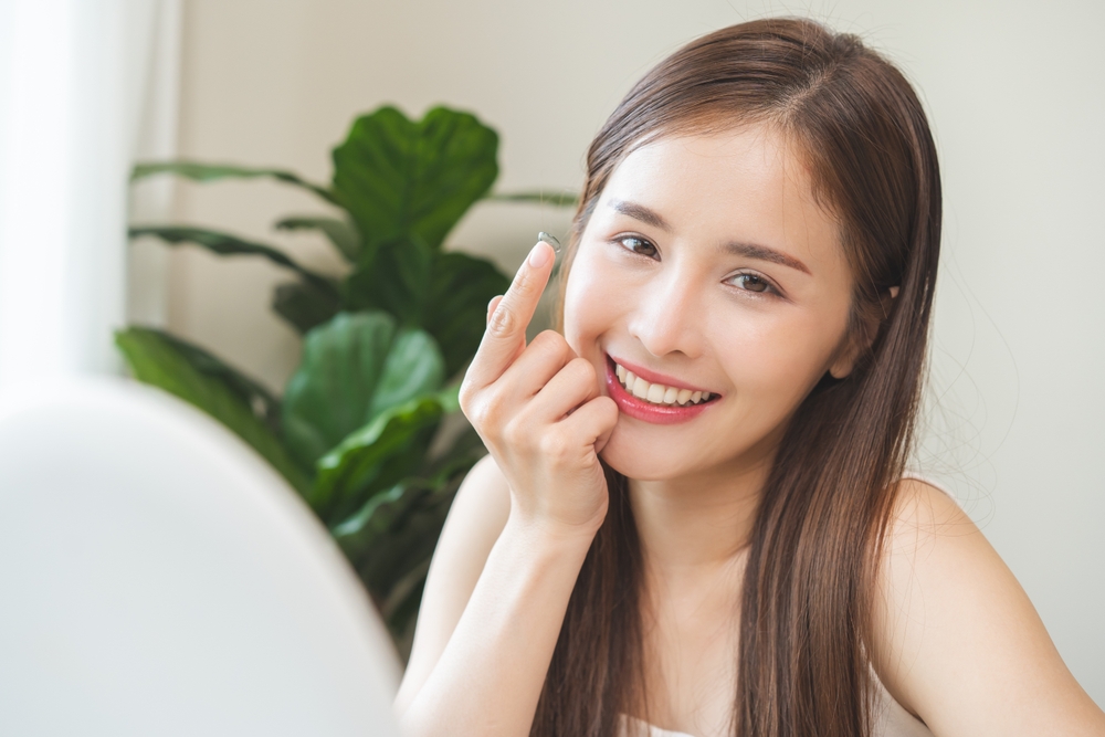 Woman Holding Contact Lens on Fingertip and Smiling A smiling young woman indoors, showing a contact lens on her fingertip while sitting near a plant and looking at the camera.