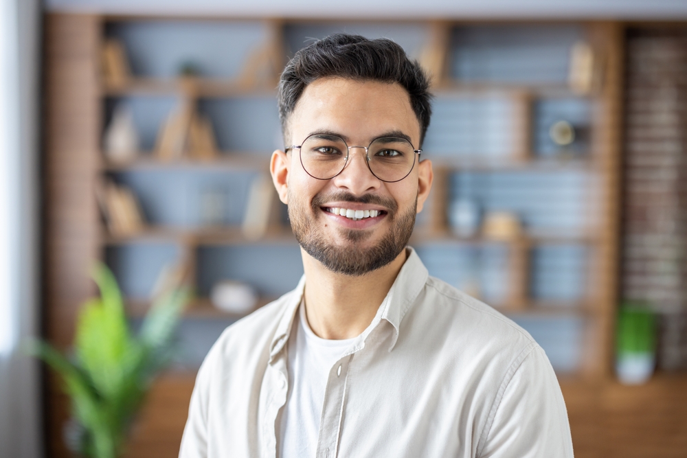 Smiling Man Wearing Glasses Indoors A young man with neatly groomed beard and glasses smiles confidently, standing in a warmly lit indoor space with shelves in the background