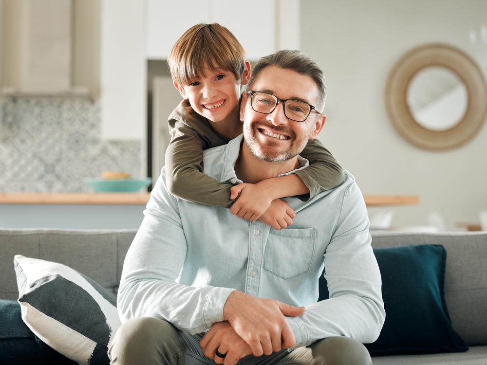A cheerful father wearing glasses sits on a couch with his young son hugging him from behind in a bright, modern living room - Eye Doctor in Colonial Heights.