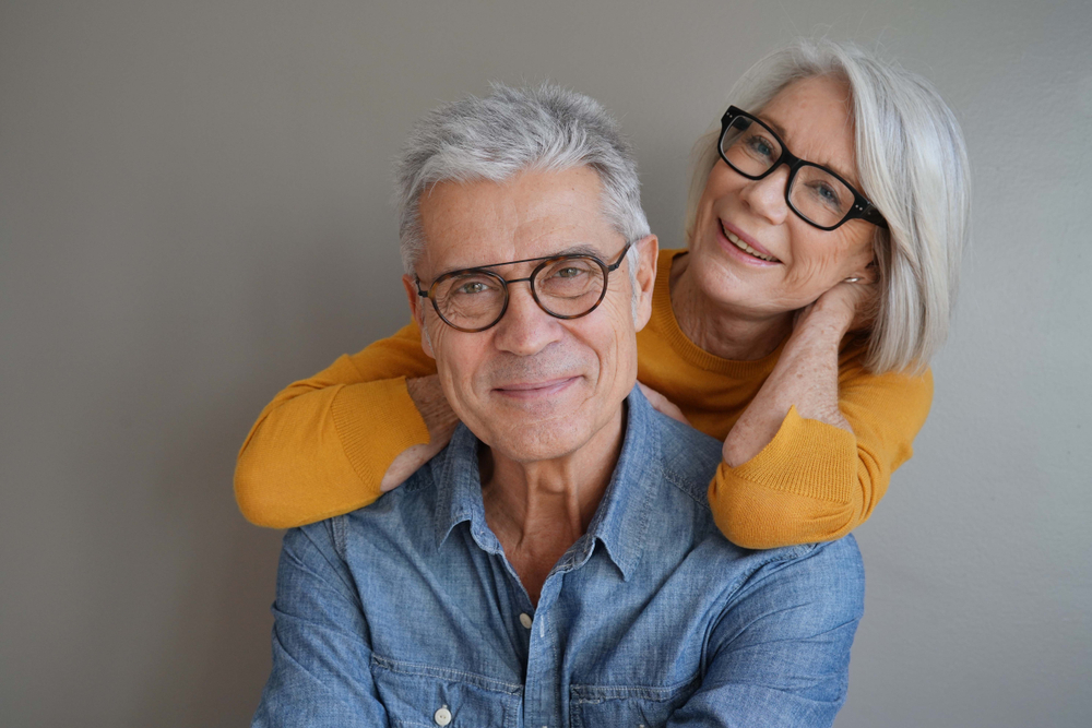 Happy Senior Couple Wearing Glasses An older couple wearing glasses smiles warmly at the camera, with the woman in a mustard sweater embracing the man in a denim shirt.