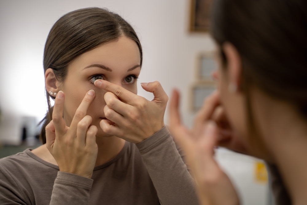 Woman Inserting Contact Lens in Front of Mirror Close-up of a woman carefully inserting a contact lens into her eye while looking in the mirror, with focused expression and gentle lighting.