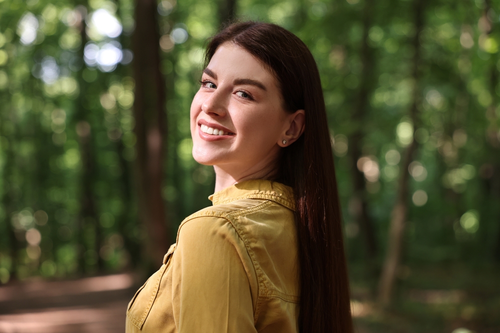 Young woman in a yellow shirt smiles while standing in a sunny wooded area, enjoying the outdoors - in Johnson City, TN.