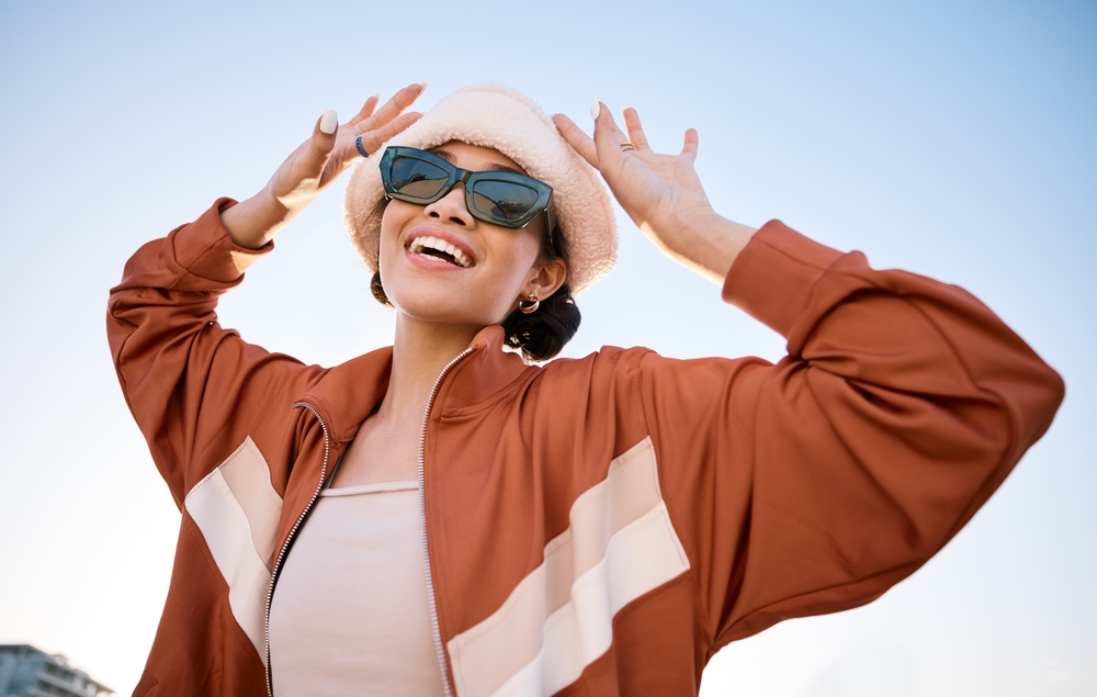 Stylish woman wearing dark sunglasses, a white beret, and a rust-colored jacket, smiling joyfully under a blue sky - Bristol Eye Doctor