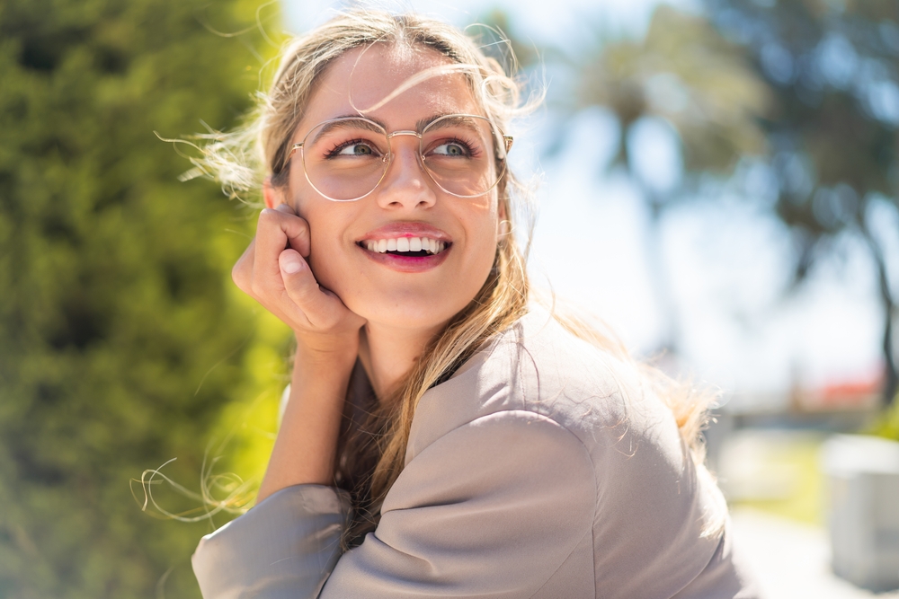 Young Woman Smiling with Glasses Outdoors A young woman with blonde hair and glasses smiles brightly while sitting outdoors on a sunny day, with trees in the background.