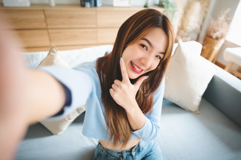 Happy Woman Taking a Selfie with Clear Vision A cheerful young woman in a light blue top taking a selfie on a couch, smiling confidently with clear eyes and natural lighting.